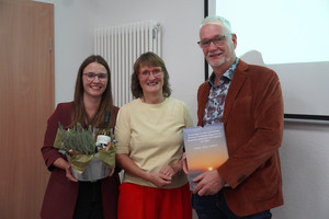 Drei Personen stehen vor einer Leinwand. Ihnen wurden Präsente überreicht.