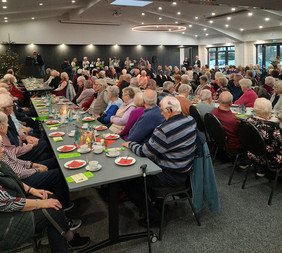 Seniorinnen und Senioren sitzen an der gedeckten Kaffeetafel und hören der Begrüßung des Bürgermeisters zu