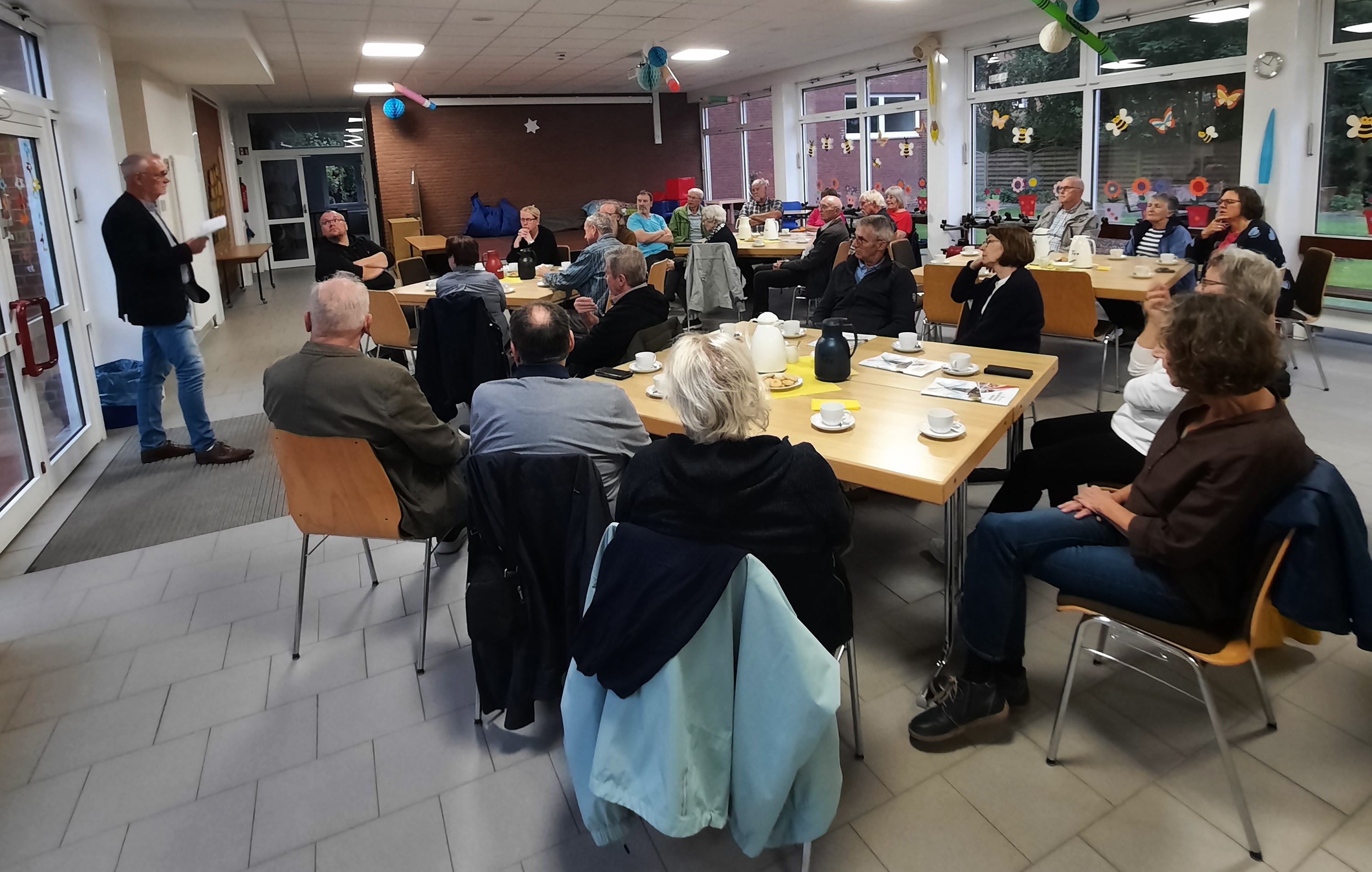 In der Pausenhalle der Grundschule Holtermoor sitzen Frauen und Männer an Gruppentischen und hören dem Bürgermeister zu.