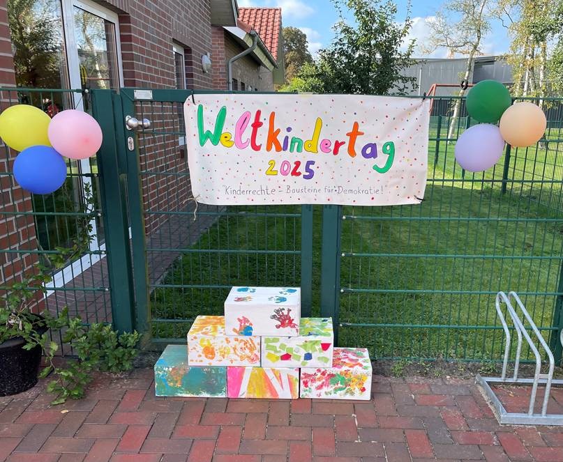 Bunt bemalte weiße Kartons stehen zu einer Pyramide vor einem Drahtzaun, darüber hängt ein buntes Plakat mit Schriftzug Weltkindertag 2025
